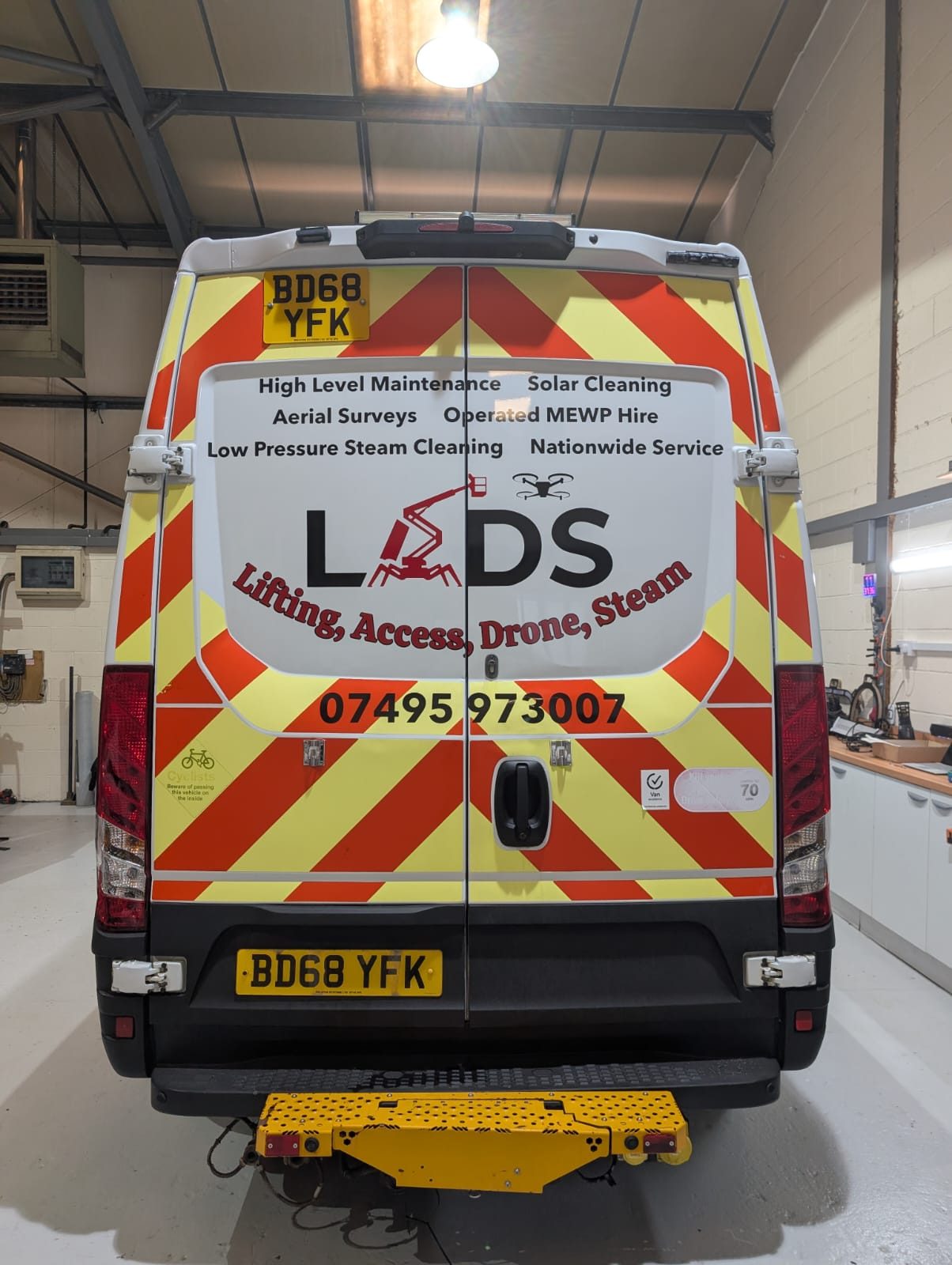 The rear of the LADS van in the workshop, its vinyl graphics listing high level maintenance, solar cleaning, aerial surveys, operated MEWP hire and low pressure steam cleaning