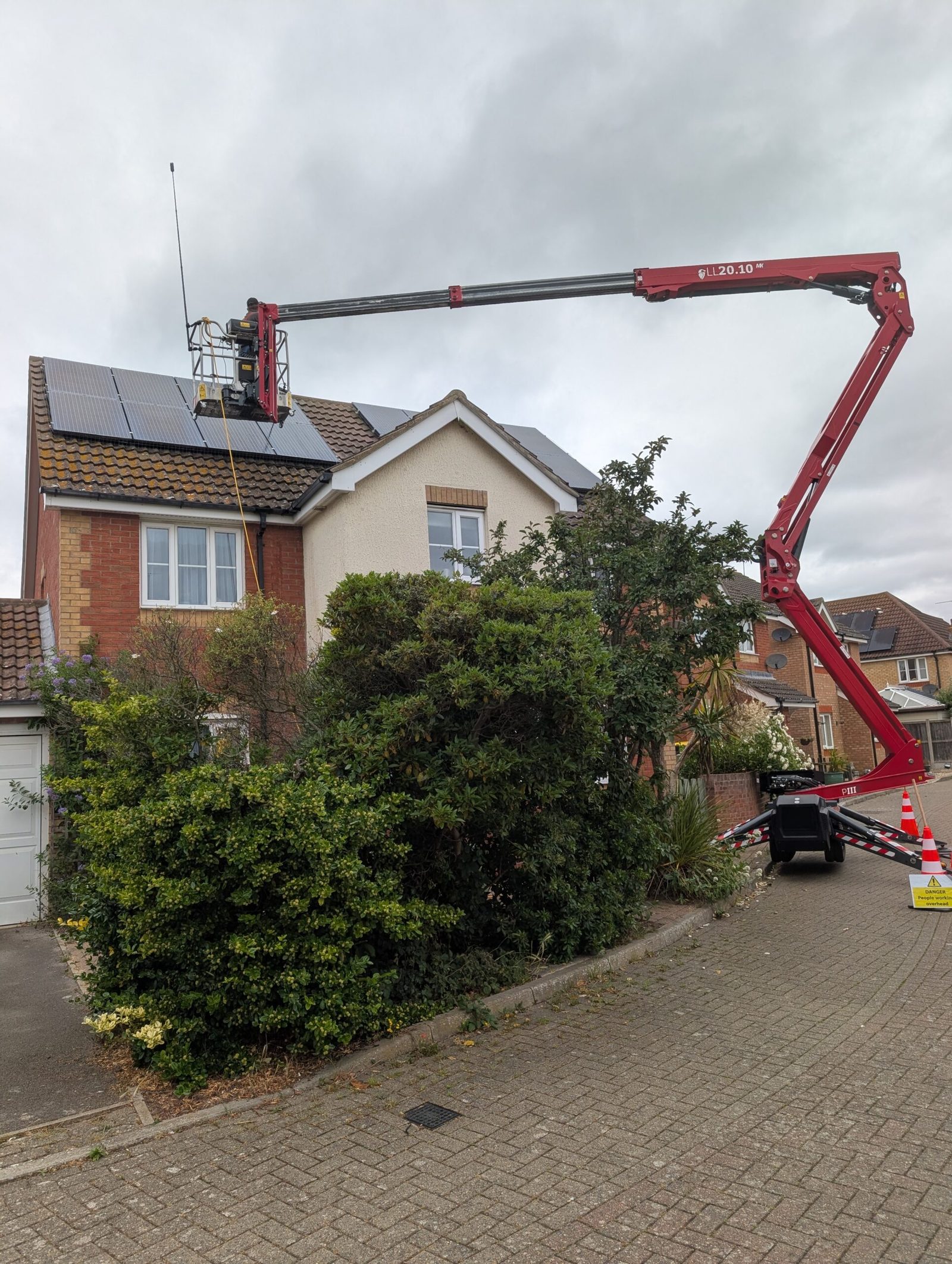 Tracked spider lift reaching over a hedge and driveway to a rooftop solar array in Essex